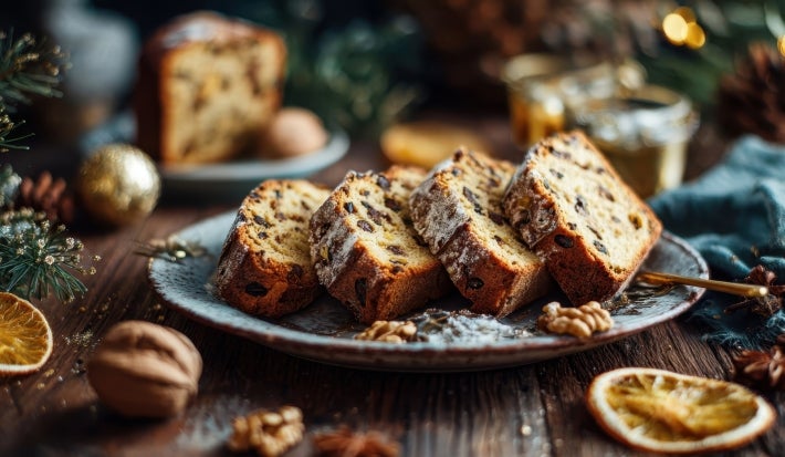Fotografia de um prato com fatias de bolo de frutas, decorado com açúcar de confeiteiro, cercado por elementos festivos como um raminho de pinheiro, nozes e fatias de laranja seca.