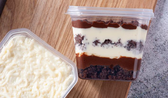 Fotografia em tons de marrom com um pote de bolo ao centro. Dentro desse pote existe camadas de um creme de cor branca e camadas de um creme de cor chocolate. Em cima do bolo existe coco ralado