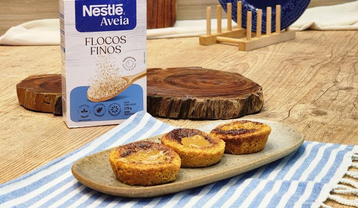 Fotografia em tons de marrom com um prato de cerâmica ao centro. Em cima do prato existe 3 muffins de doce de leite posicionados lado a lado. Ao fundo existe uma embalagem de Aveia Flocos Finos.