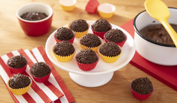 Fotografia em tons de vermelho e amarelo em uma bancada de madeira, um guardanapo de pano vermelho e branco listrado, uma tábua de madeira vermelha, um suporte para bolo branco com vários brigadeiros em forminhas amarelas e vermelhas em cima dele. Fotografia em tons de vermelho e amarelo em uma bancada de madeira, um guardanapo de pano vermelho e branco listrado, uma tábua de madeira vermelha, um suporte para bolo branco com vários brigadeiros em forminhas amarelas e vermelhas em cima dele.