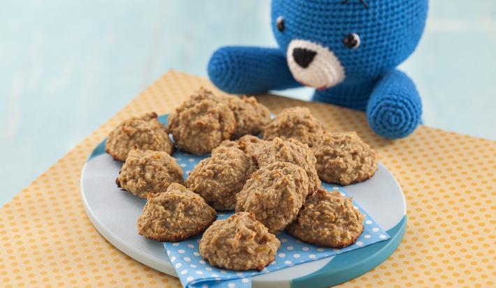 Fotografia em tons de marrom em uma bancada de madeira de cor laranja. Ao centro, há um prato azul contendo os cookies e ao lado, há um ursinho azul sentado.