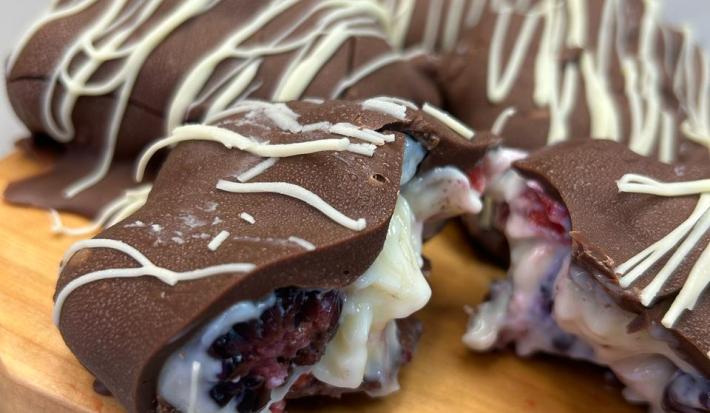 Fotografia de uma tábua de madeira clara sobre uma mesa cinza claro. Sobre a tábua tem alguns bombons coobertos com chocolate meio amargo e decorados com chocolate branco, o recheio é um creme de ninho com frutas vermelhas, um deles está aberto.
