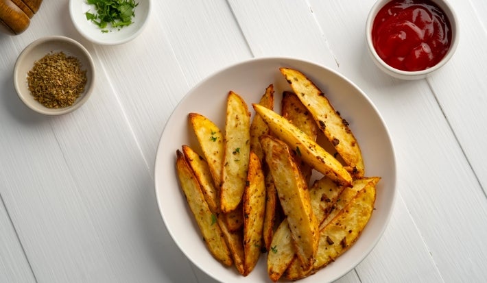 Fotografia em tons claros de um recipiente branco médio, fundo, sobre uma mesa de madeira branca. Dentro do recipiente tem batatas assadas e temperadas, cortadas em meia lua. Ao lado do recipiente tem temperos e ketchup em potinhos brancos.