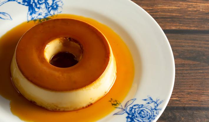 Fotografia de um prato branco, com desenhos azuis, grande raso sobre uma mesa de madeira. Sobre o prato tem um pudim pequeno tradicional feito com amido de milho na receita.