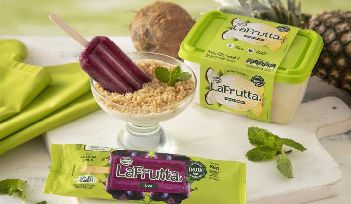 Foto da receita de Taça Refrescante de Sorvete LAFRUTTA Abacaxi + Coco com Farofa Crocante, servida numa taça decorada com picolé de uva, sobre uma bancada clara com um pano verde claro, um coco fresco e um abacaxi inteiro.