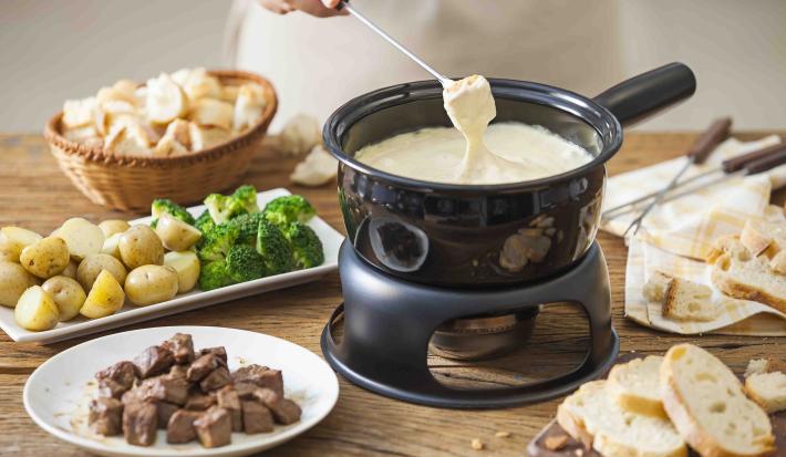 Fotografia em tons de marrom e beje de uma bancada de madeira com uma panela de fondue preta com fondue de queijo. Ao lado um prato redondo com carne, um prato retangular com batatas e brócolis, e uma cesta com pães.