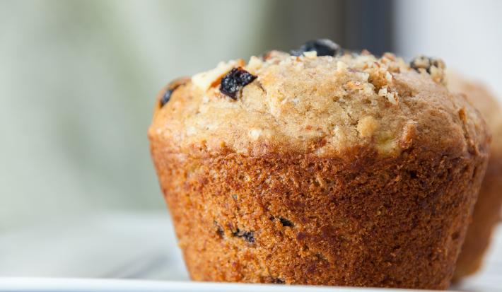 Fotografia em tons claros de um muffin de passas e aveia sobre um apoio branco.