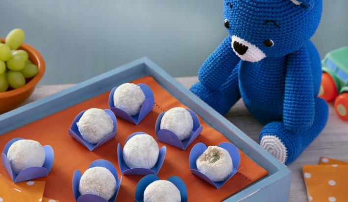 Fotografia em tons de branco em uma bancada de madeira de cor branca. Ao centro, uma mini mesa azul com uma toalha laranja em cima e dentro, há os docinhos. Ao lado, está o ursinho azul e ao fundo, há um carrinho e um pote de uvas verdes.