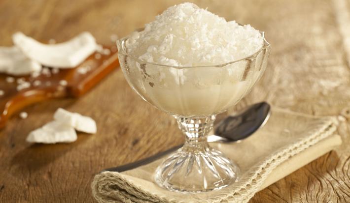 Fotografia em tons de marrom de uma bancada marrom, sobre ela um paninho beje, um taça com raspadinha e uma colher. Ao fundo uma tábua com pedaços de coco.