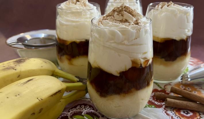 Foto da receita de banana cremosa servida em 4 porções em copos largos de vidro, sobre uma mesa decorada com uma toalha colorida, algumas bananas em cacho e paus de canela. Ao fundo um bowl de porcelana.