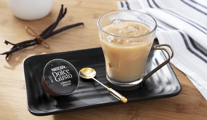 Foto em tons de bege da receita de espresso com baunilha e leite gelado servida em uma xícara de vidro sobre uma base de porcelana preta com uma colher dourada e uma cápsula de dolce gusto espresso intenso
