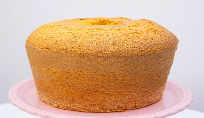 Um bolo de cenoura assado em forma redonda com buraco no meio, colocado sobre um prato rosa. O bolo está dourado por fora, com a superfície levemente rachada, indicando que está bem fofinho.