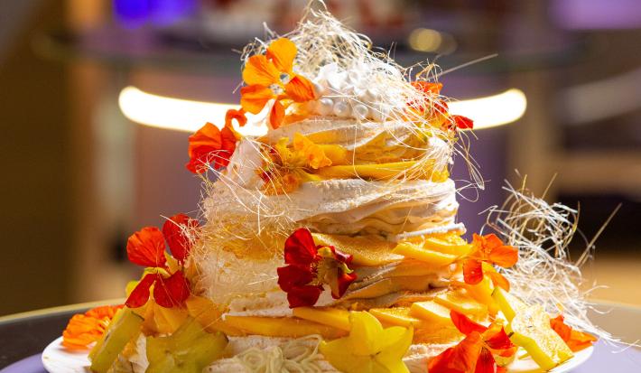 Foto aproximada da receita de Pavlova Tropical, lindíssima e montada em camadas de massa com recheio e decorada com flores comestíveis e arabescos de chocolate branco