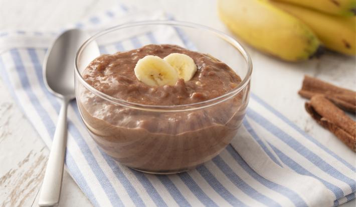 Fotografia em tons de azul em uma bancada de madeira clara com um paninho listrado azul e em cima um potinho de vidro com o doce de banana e cacau. Ao lado, algumas bananas, uma colher e canela em pau.