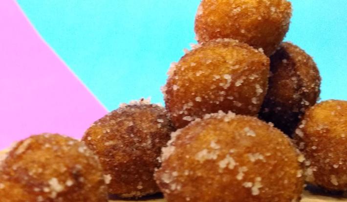 Fotografia de vários bolinhos doces fritos passados no açúcar e canela. Os bolinhos estão juntos, sobre uma tábua de madeira, com um fundo rosa e azul claro forte.