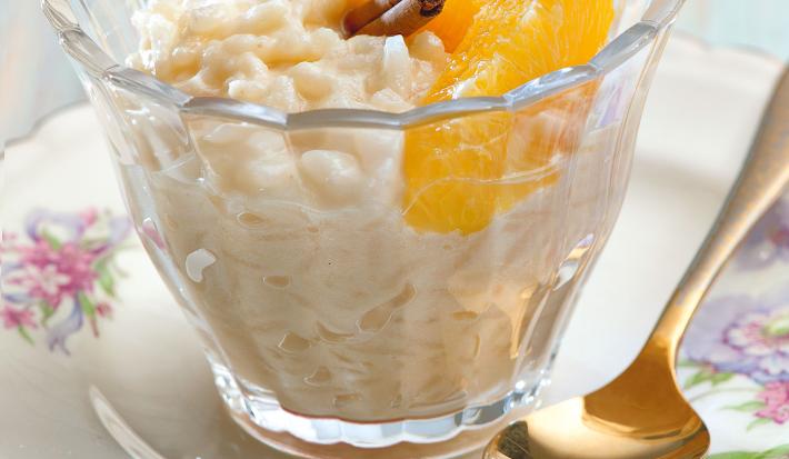 fotografia em tons de branco e amarelo tirada de um prato redondo branco com um copo transparente que contém arroz doce com rodelas de laranja de canela
