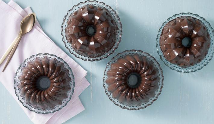 Fotografia em tons de chocolate, azul e rosa, com quatro porções de pudim de chocolate em pratinhos de cristal, um deles sobre guardanapo rosa tendo ao lado colheres de sobremesa, tudo sobre bancada azul.