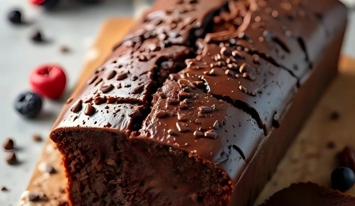 Bolo de chocolate cremoso em uma tábua de madeira, com cobertura brilhante e raspas de chocolate. Uma fatia foi cortada, revelando um interior denso.