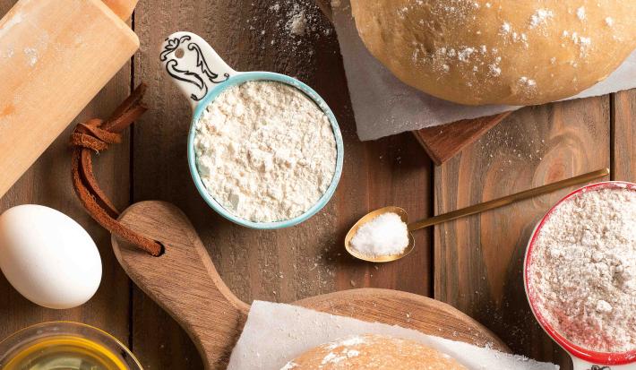 Fotografia em tons de branco e marrom, com massas para macarrão sobre papel manteiga e peças de madeira, no entorno potinho com ovo. ovo inteiro, potinhos com farinha de trigo, colher com sal, rolo de abrir massa, tudo sobre bancada de madeira.