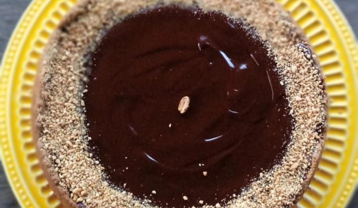 Foto da receita de Torta de Amendoim. Observa-se uma torta com ganache e farofa de amendoim em cima sobre um prato de amendoim.