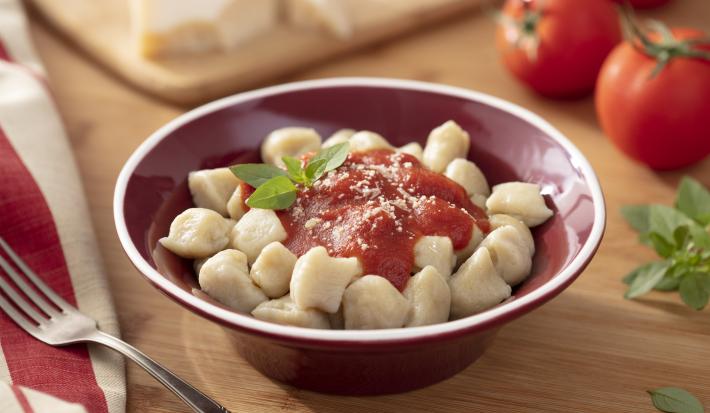 Imagem de um bowl vermelho com nhoque de mandioca com molho de tomate. Ao fundo, tomates, manjericão e um pedaço de queijo parmesão.