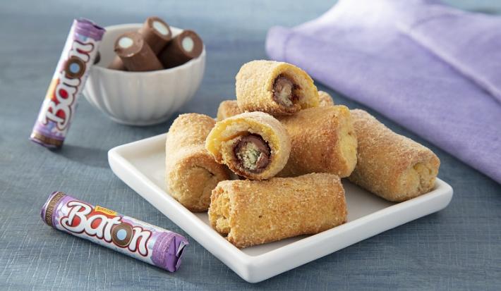Foto da receita de rolinhos de baton algodão doce servida em diversas porções sobre uma base de porcelana quadrada em cima de uma mesa acinzentada com um paninho lilás e algumas unidades de baton ao lado