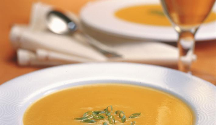 Fotografia em tons de laranja em uma mesa de madeira, com uma taça de vinho branco e um prato branco fundo com a sopa de abóbora, maçã e gengibre dentro dele.