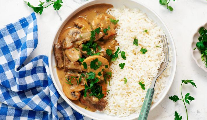 Imagem vista de cima de um prato branco com arroz e strogonoff de carne bem cremoso. Ao lado, um paninho xadrez azul e folhas de salsinha decorando.