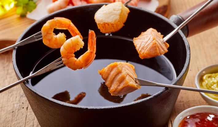 Fotografia em tons claros com uma panela de fondue ao centro. Alguns pedaços de frutos do mar estão espetados no palito para serem mergulhados no fondue