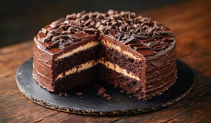 Em cima de uma mesa, prato redondo com bolo de chocolate fofo, recheado com creme branco, cobertura de chocolate e chocolate ralado.