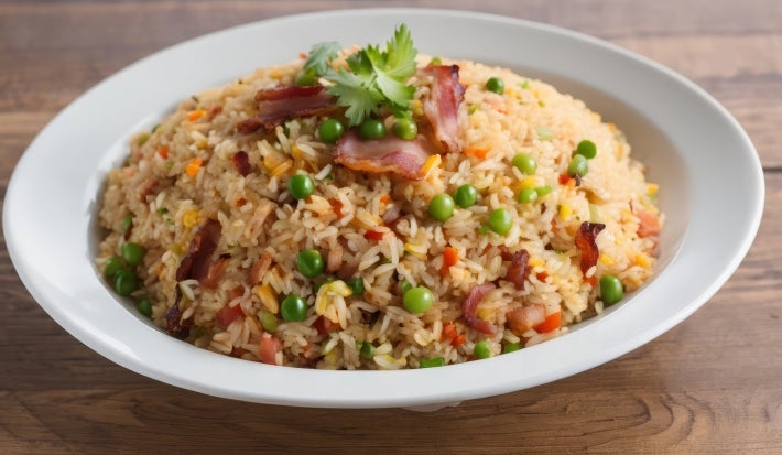 Em cima de uma mesa, prato redondo com porção de arroz soltinho com ervilhas, cenouras picadas, pedaços de bacon, decorado com fatias de bacon e folha de salsa.