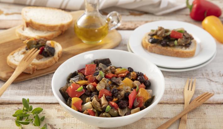Fotografia em tons de vermelho e amarelo em uma bancada de madeira clara, um prato redondo branco grande ao centro com a caponata de berinjela. Ao lado, uma tábua de madeira com fatias de pães italianos, um potinho de azeite e pimentões amarelo e vermelho