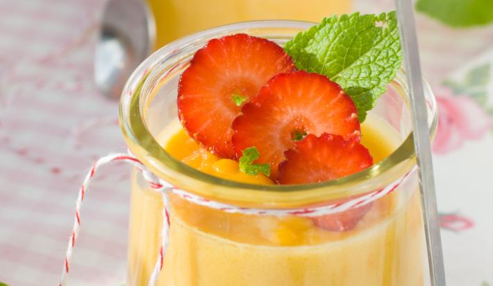 Fotografia de dois recipientes de vidro decorativos com uma mousse de laranja, no recipiente da frente, em cima da mousse, há três fatias de morangos com hortelã ao lado. Ao fundo, no recipiente há apenas a mousse de laranja.