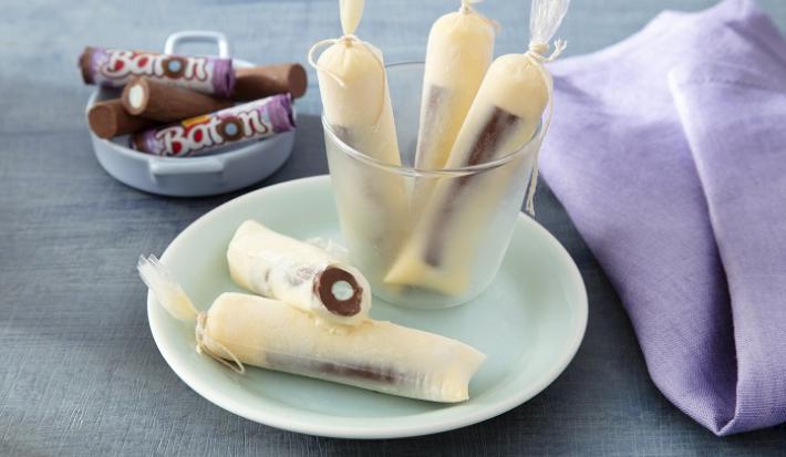 Foto da receita de geladinho algodão doce servida em 5 porções dentro de um pote transparente em cima de um prato de porcelana com um paninho lilás e algumas unidades de baton ao fundo