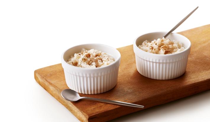 Foto da receita de Arroz Doce Vegano. Observa-se uma tábua de madeira com dois potinhos brancos e pequenos de arroz doce em cima.