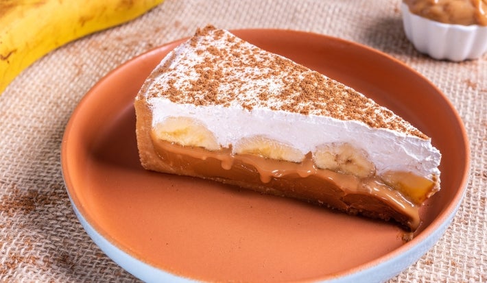 Em cima de uma mesa, prato redondo com fatia de banoffee com massa de biscoitos crocante, doce de leite, bananas em rodelas, decorado com chantilly cremoso e polvilhado com canela em pó.