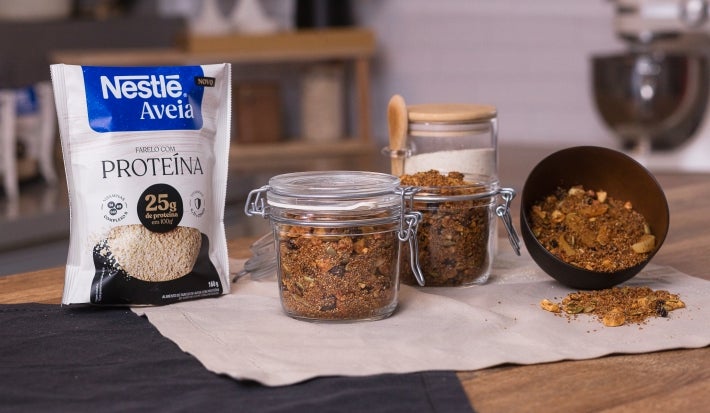 Foto da receita de granola proteica com aveia servida em dois potes herméticos de vidro sobre um pano neutro em cima de uma mesa de madeira com uma embalagem de aveia proteína ao lado