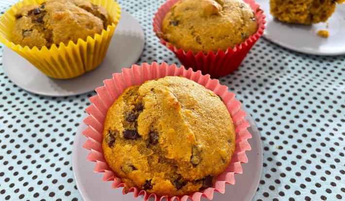 Fotografia de três forminhas de cupcake com bolo de cenoura e uva passa, sendo duas de tom rosa e uma amarela. Duas delas estão sobre um miniprato cinza raso, os quais estão sobre uma toalha de mesa de tom azul claro com bolinhas pretas.