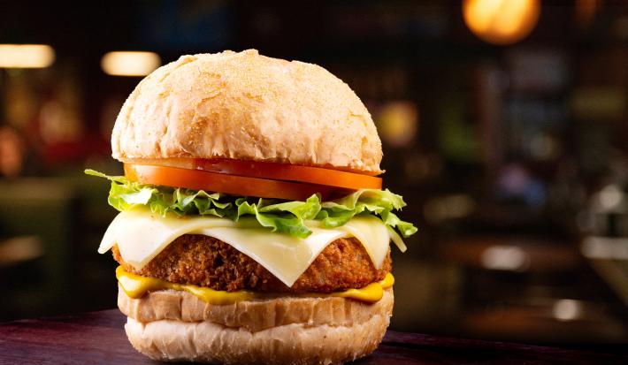 Fotografia de um hamburguer com tomate, alface, queijo, hambúrguer empanado e um molho amarelo situado ao centro de uma foto em tons escuros