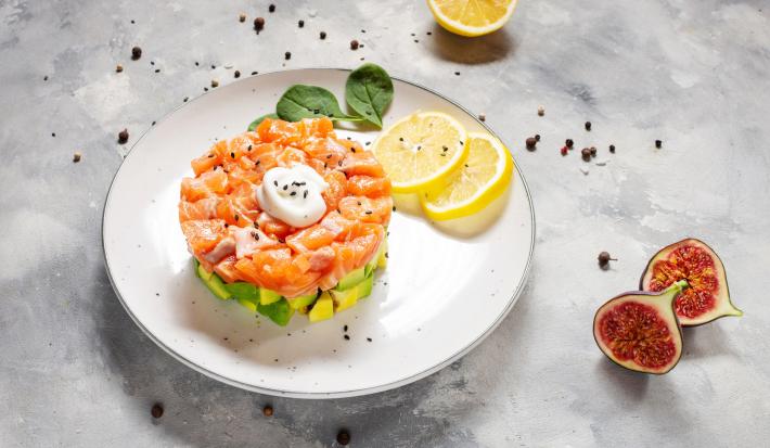 Fotografia em tons claros de uma mesa de cimento queimado claro. Sobre a mesa tem um prato branco raso com tartar de salmão sobre abacate em cubos ao lado de duas rodelas de limão siciliano.