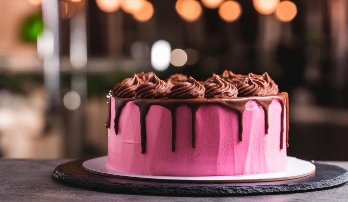 Fotografia de um bolo redondo coberto com um vibrante glacê, adornado com ganache de chocolate brilhante que escorre pelas laterais. O fundo desfocado com luzes quentes cria uma atmosfera acolhedora.
