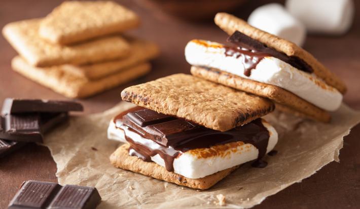 Fotografia de duas peças de s'mores sobre um papel manteiga. Ao fundo tem alguns pedaços de chocolate e alguns biscoitos empilhados, sobre uma mesa de madeira.