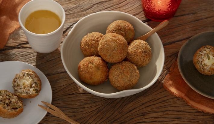 Foto em tons de laranja da receita de arancini de abobrinha servida em diversas porções em um bowl de cerâmica com uma colher de madeira sobre uma mesa de madeira. Ao lado há mais dois pratos com arancini e um copo de suco de laranja