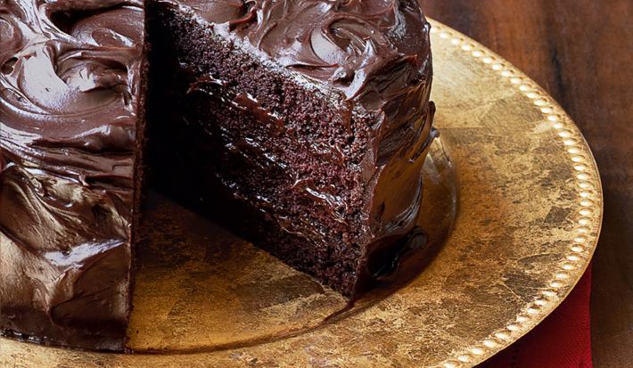 Fotografia de um bolo marrom de chocolate com uma fatia faltando, em um prato dourado em cima de um guardanapo de tecido vermelho em uma mesa de madeira escura.