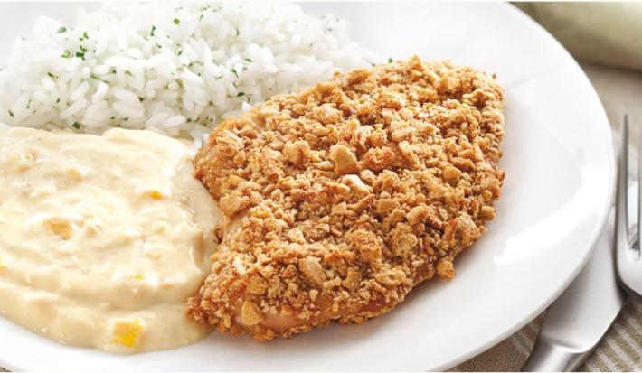 Fotografia em tons de branco, amarelo e dourado, ao centro prato branco contendo creme de milho, arroz e frango empanado, ao lado uma faca e guardanapo cinza, sobre toalha listrada em tons ocre.