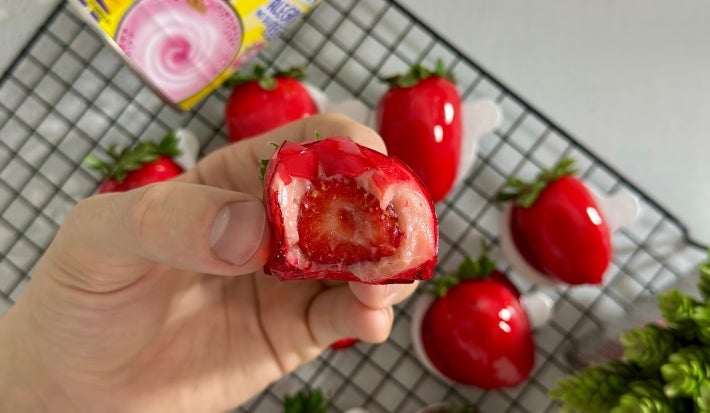 Fotografia mosta uma mão segurando um o doce com morango coberto por brigadeiro de Nesquik e calda vermelha brilhante, no centro da foto. Ao fundo, outros doces aparecem em desfoque.