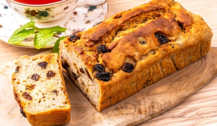 Fotografia mostra um pão de passas recém-assado, parcialmente fatiado, exibe uma crosta dourada e interior macio. Ao lado, uma xícara de chá vermelho em um pires floral, com um raminho de hortelã