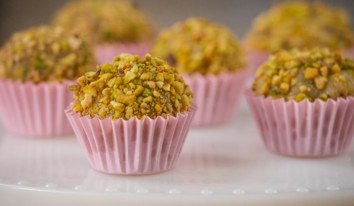 Fotografia de seis forminhas de cor rosa com brigadeiro de pistache enrolado no pistache triturado. Os brigadeiros estão em um apoio branco decorativo.