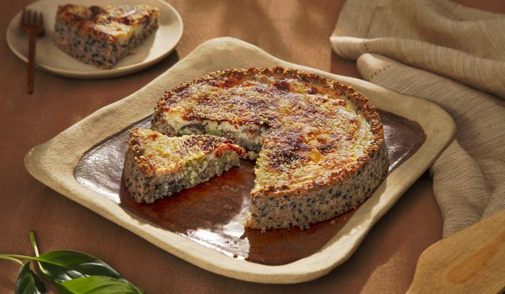 Foto da receita de quiche de aveia corte rústico servido em uma porção com uma fatia cortada sobre uma base quadrada de cerãmica marrom com um pano de linho bege ao lado