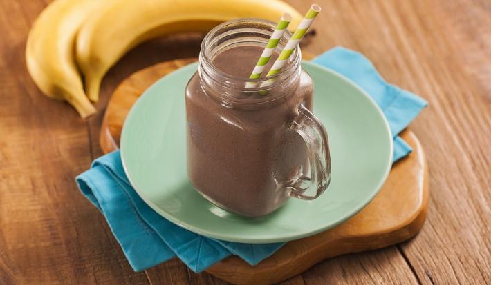 Fotografia em tons escuros, em uma bancada de madeira de cor marrom. Ao centro, uma tábua de madeira marrom contendo um pano azul. Em cima, há um prato verde e redondo contendo um copo com o milkshake. Ao fundo, há 2 bananas.